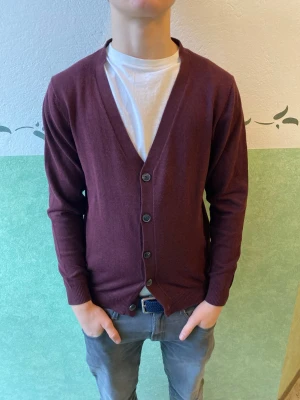 Vinröd cardigan från Jack and Jones  - Vinröd cardigan med knappar från Peter England. Klassisk V-ringning och ribbade muddar vid ärmslut och nederkant. Perfekt att slänga över en t-shirt för en chill look. Materialet är mjukt och tunt, vilket gör den enkel att bära och styla.