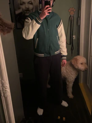 Karl Kani Jacka - Vinted College Bombarjacka Dark/Olive Green - Superfin och trendig & vintage varsityjacka från Karl Kani i klassisk streetwear‑stil. Jackan har en grön kropp, cremefärgade ärmar och tydliga KANI‑detaljer på ärm och rygg. Perfekt för dig som gillar en sportig och urban look.  Skick: Mycket bra – sparsamt använd och väl omhändertagen. Storlek: (Medium; M)  Material: 70% polyester, 30% ull Ärmar: 100% polyester Foder: 100% polyester  Detaljer: Rundad hals och ståkrage Knappstängning Flerfärgat mönster Isticksfickor Midjeresår