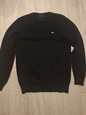 Svart stickad tröja Lyle & Scott - Klassisk svart stickad tröja från Lyle & Scott med rund hals och den ikoniska gula logon broderad på bröstet. Tröjan har långa ärmar och ribbade muddar vid ärmslut och nederkant. Perfekt för en stilren och enkel look.
