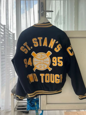Blå och gul collegejacka  - Vintage collegesjacka i mörkblå med gula detaljer och broderade texter. Jackan har tryckknappar, ribbade muddar och krage med gula ränder. Stor brodyr på ryggen med basebolltema och årtal, samt namn broderat fram. 