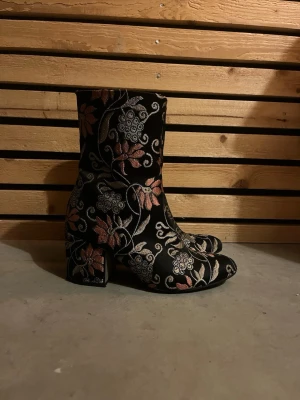 Blommiga svarta stövletter med klack - Unika svarta stövletter med broderade blommor i rosa, silver och beige. Skorna har en fyrkantig klack och rundad tå. Materialet är textil med tydlig struktur och mönster. Perfekta för dig som vill sticka ut med din stil.