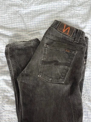 Grim Tim, Dry black selvage jeans från Nudie Jeans 30/34 - Säljer ett par svarta jeans från Nudie Jeans med klassisk femficksmodell och raka ben. Jeansen har en snygg tvättad look och detaljer i orange, inklusive loggan på bakfickan och läderpatchen i midjan. Tillverkade i bomull för skön passform. (W30 L34)