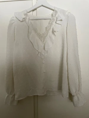 Vit blus med volang och spetsdetaljer - Romantisk vit blus med volang längs V-ringningen och spetsdetaljer. Blusen har diskreta prickar i tyget, knappar framtill och långa ärmar med volang vid ärmslut. Perfekt för en söt och feminin look.