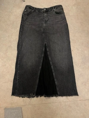 Svart jeanskjol med slits - Snygg svart lång jeanskjol med hög midja och rå kant nertill. Kjolen har en tydlig slits framtill som ger en cool look. Klassisk femficksmodell och knappgylf. Perfekt för dig som gillar streetstyle.