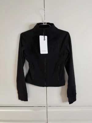 Svart croppad träningsjacka Lululemon - Snygg svart croppad jacka från Lululemon i mjukt och stretchigt Nulu-material med inslag av Lycra. Jackan har hög krage, långa ärmar med tumhål, två dragkedjeförsedda fickor och slim fit. Perfekt för träning eller som ett sportigt lager.