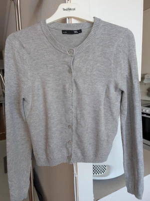 Ljusgrå kofta Lager 157 S - Ljusgrå, tunnstickad kofta från Lager 157 i stlk S med rund halsringning och sju knappar framtill. Klassisk och enkel design med långa ärmar och ribbade muddar vid ärmslut och nederkant. Perfekt att slänga över en t-shirt eller linne.