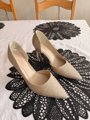Glittriga guldiga pumps från Tiamo - Säljer ett par glittriga pumps från Tiamo i guldigt tyg. Skorna har spetsig tå, öppen insida och smal klack. Klacken är 8cm hög.