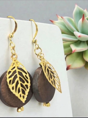 Guldörhängen med trä och lövdetalj - Unika örhängen med rund mörk träplatta och dekorativa guldfärgade metallblad. Krokarna är guldpläterade. Hängande modell med snygg kontrast mellan det naturliga träet och det blanka guldet. Perfekta för dig som gillar boho och naturinspirerad stil.