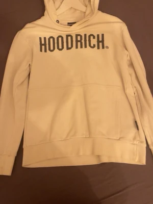 Beige hoodie från Hoodrich - Snygg beige hoodie från Hoodrich med stor svart logga på bröstet. Klassisk huva och stor magficka framtill. Perfekt för en avslappnad streetwear-look. Mjuk och skön bomullsblandning, ribbade muddar vid ärmar och nederkant.