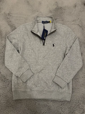 Grå halvzip sweatshirt från Polo Ralph Lauren - Snygg grå sweatshirt med halv dragkedja från Polo Ralph Lauren. Tröjan har ribbade muddar, hög krage och den klassiska broderade loggan på bröstet. Perfekt för en clean och avslappnad stil. Tillverkad i mjuk bomullsblandning. Står storlek M men skulle nog säga tröjan passar bättre på Xs/S