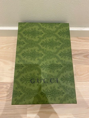 Box  - Säljer en snygg grön presentbox med elegant mönster och logga på locket. Perfekt för att förvara eller ge bort exklusiva accessoarer. Boxen har en lyxig känsla och sticker ut med sin unika design.