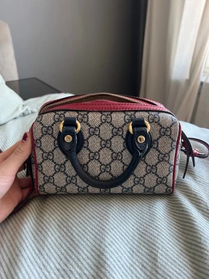 Gucci beige monogram handväska - Säljer en snygg beige handväska från Gucci med klassiskt GG-monogram, mörkblå handtag och detaljer samt röda kanter och axelrem. Väskan har guldfärgade metalldetaljer och dragkedja upptill. Köpt från hemsidan A Retro tale för 8000kr, pris kan diskuteras.