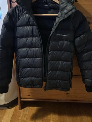 Svart pufferjacka Peak Performance - Snygg svart pufferjacka från Peak Performance med huva och dragkedja framtill. Jackan har quiltad design och är perfekt för kyliga dagar. Klassisk passform och diskret logga på bröstet ger en stilren look. Inga hål