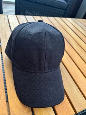 Svart klassisk keps i bomull - En stilren svart keps med böjd skärm och sex paneler. Kepsen har ventilationshål och är tillverkad i ett mjukt bomullstyg. Perfekt för dig som vill ha en enkel och tidlös look.