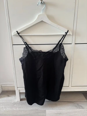 Svart spetslinnetopp från ONLY - Säljer en svart linnetopp från ONLY med tunna axelband och vacker spetsdetalj vid urringningen. Toppen har en enkel och stilren design som passar perfekt till många olika outfits. Materialet känns mjukt och lätt mot huden.