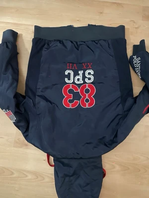 Mörkblå vindjacka från Santoz Polo Club - Stilren mörkblå vindjacka från Santoz Polo Club med röda och vita detaljer. Stor broderad 83 på ryggen, broderade loggor på ärmen och insidan har röd quiltad lining med stort klubbmärke. Jackan har huva och muddar vid ärmslut och midja.