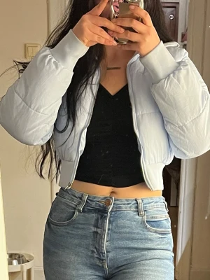 Ljusblå cropped pufferjacka - Säljer en ljusblå cropped pufferjacka med dragkedja framtill och breda muddar vid ärmslut och midja. Jackan har en boxig passform och quiltad design som ger en trendig look. Perfekt för dig som vill ha en snygg och varm jacka till vintern.