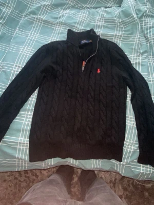 Svart kabelstickad tröja  - Svart kabelstickad tröja med halv dragkedja och hög krage. Tröjan har det klassiska röda broderad på bröstet och ribbade muddar vid ärmslut och nederkant. Perfekt för en stilren och avslappnad look.