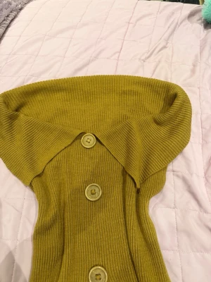 Olivgrön ribbstickad kofta med krage - Säljer en olivgrön ribbstickad kofta med bred krage och stora matchande knappar framtill. Koftan har en figurnära passform och är perfekt för lager på lager. Materialet känns mjukt och stretchigt, vilket gör den både bekväm och snygg.