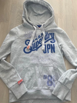 Ljusgrå hoodie från Superdry - Snygg ljusgrå hoodie från Superdry med blått tryck och japanska tecken på bröstet. Klassisk känguruficka framtill, vita dragsnören i huvan och ribbade muddar. Perfekt för en avslappnad streetwear-look.