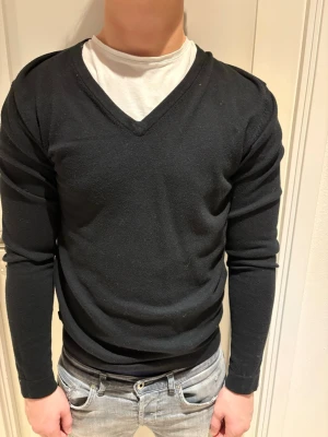 Svart stickad V-ringad tröja - Klassisk svart stickad tröja med V-ringning och långa ärmar. Tröjan har en enkel och stilren design utan synliga loggor eller mönster. Passar perfekt till jeans eller över en t-shirt för en avslappnad look.