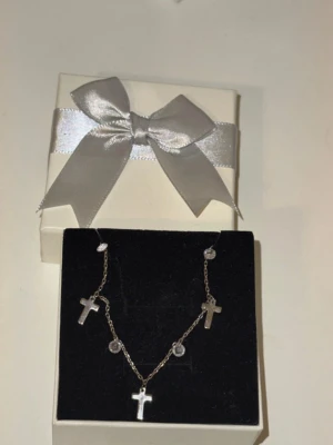 Silverhalsband med kors och stenar - Snyggt halsband i äkta silver (stämplat 925) med tre små korshängen och runda klara stenar på en tunn kedja. Kommer i en elegant presentask med silverfärgat band. Perfekt för dig som gillar stilrena och trendiga smycken.