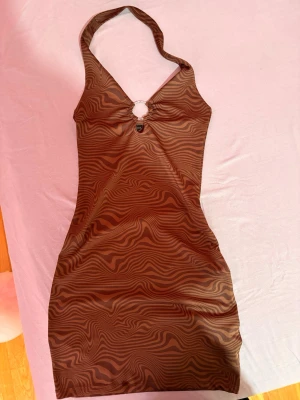 Brun mönstrad halterneckklänning - Brun kort halterneckklänning med ringdetalj framtill. Klänningen har en tight passform och är ärmlös. Endast provat (i bilden) Perfekt till våren/sommaren. PM om ni har frågor ❤️