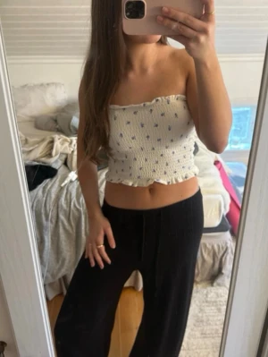 Vit blommig bandeau tubtopp - Supersöt bandeau tubtopp i vit med små blå blommor. Toppen är smockad och har volangkant både upptill och nedtill. Perfekt för varma dagar och ger en fräsch och somrig vibe.