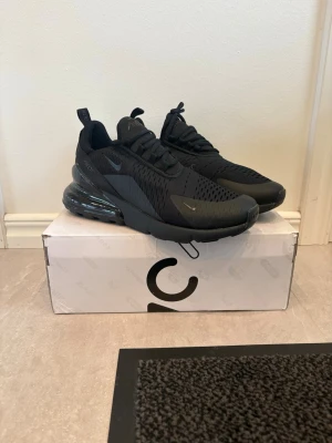 Nike Air Max 270 helsvarta sneakers - Snygga helsvarta Nike Air Max 270 sneakers med mesh- och syntetmaterial, ikonisk synlig Air-enhet i hälen och diskreta loggor på sidan och plösen. Skorna har en sportig siluett och är perfekta för dig som gillar streetwear och komfort.