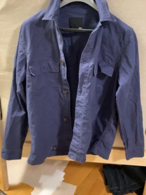 Blå overshirt med knappar - Stilren blå overshirt med klassisk krage och knäppning framtill. Skjortan har två bröstfickor med lock och långa ärmar. Perfekt att bära öppen eller stängd för en avslappnad look. Pris går jobba 