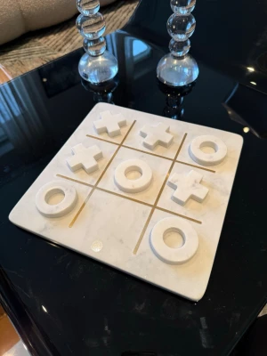 Tic Tac Toe i vit marmor - Snyggt Tic Tac Toe-spel i vit marmor med guldfärgade linjer. Spelplanen är fyrkantig och brickorna består av kryss och ringar, även de i vit marmor. Perfekt som inredningsdetalj eller för att spela med vänner. Ger ett lyxigt och modernt intryck.Gjord av Lene Bjerre 
