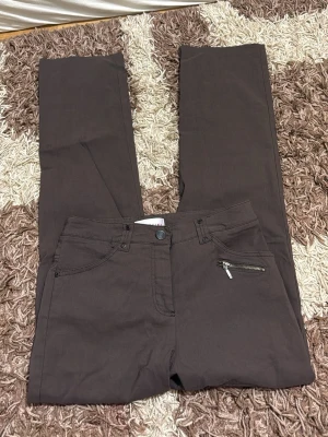 Bruna raka jeans med dragkedjedetalj - Snygga bruna jeans med rak passform och en cool dragkedjedetalj på fickan framtill. Klassisk femficksmodell och knappgylf. Perfekta för dig som vill ha en stilren men unik look.