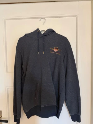 Mörkblå hoodie från GANT - Mörkblå/grå hoodie från GANT med broderad logga och text på bröstet. Klassisk känguruficka framtill, justerbar huva med snören och ribbade muddar. Perfekt för en avslappnad och sportig stil. Materialet känns mjukt och bekvämt.