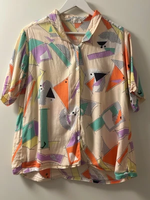 Färgglad retro skjorta med grafiskt mönster - Unik vintage skjorta med kort ärm och knappar framtill. Skjortan har ett färgstarkt grafiskt mönster. Perfekt för dig som gillar statement-plagg och vill sticka ut. Står att det är storlek L men passar mer som en M