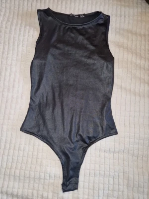 Svart glansig body från Boohoo - Snygg svart body från Boohoo i glansigt material. Rund halsringning och hög benskärning. Knäppning vid grenen,Perfekt att styla med jeans eller kjol för en trendig look.🌞😍 oandvänd