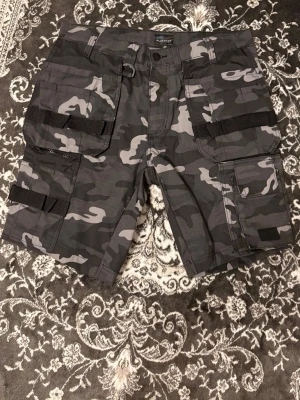 Camouflage shorts från Bluewear - Snygga shorts från Bluewear i grått och svart camouflage-mönster. De har flera praktiska fickor, bälteshällor och detaljer med svarta band. Perfekta för dig som gillar en tuff och funktionell stil.