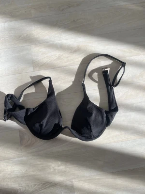 Svart bikini  med tunna axelband - Snygg svart bikini med bygel och tunna, justerbara axelband. Kuporna är lätt vadderade och har en slät, diskret ribbad struktur. Klassisk modell utan synligt märke, perfekt för dig som vill ha en enkel och stilren bh.
