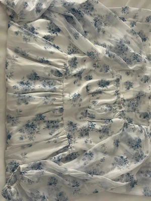 Vit blommig klänning med puffärm - Romantisk vit klänning med blått blommönster och puffiga, transparenta ärmar. Klänningen har en markerad midja och en luftig kjol som ger en söt och somrig look. Perfekt för dig som gillar en feminin stil med vintagevibbar.