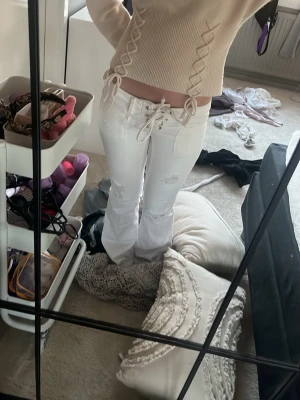 Vita snörade jeans med slitningar - Säljer ett par vita jeans med snörning i midjan och på sidorna. Jeansen har trendiga slitningar på benen och en rak passform. Perfekta för dig som vill sticka ut med detaljer och gillar en edgy stil. Använt dem två gånger så dem är inte slitna alls