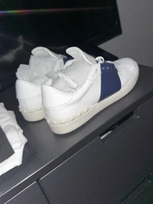 Vita sneakers med blå detalj Valentino - Säljer ett par snygga vita sneakers från Valentino med en bred marinblå rem över mitten och ikoniska nitar på hälen. Skorna har klassisk snörning och är tillverkade i skinn för en clean och lyxig look. Använda ett fåtal gånger!