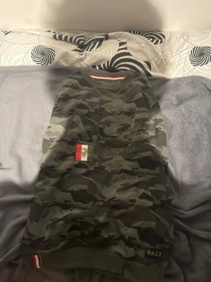 Camo sweatshirt från A.C.E - Cool camouflagemönstrad sweatshirt från A.C.E med tryck på bröstet och broderad detalj i rött, vitt och svart. Tröjan har rund halsringning, långa ärmar och ribbade muddar. Perfekt för en streetwear-look. Sprillans ny aldrig använd. 