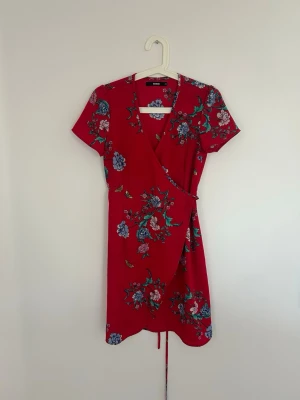 Röd blommig omlottklänning Bik Bok - Snygg röd omlottklänning från Bik Bok med blommigt mönster i blått, vitt och grönt. Klänningen har korta ärmar, v-ringning och knytband i midjan. Perfekt för sommarens alla tillfällen!