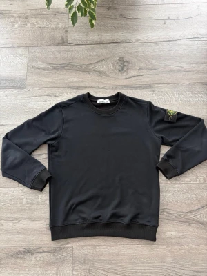 Svart sweatshirt från Stone Island - Klassisk svart sweatshirt från Stone Island med rund hals och ribbade muddar. Tröjan har den ikoniska Stone Island-loggan på vänster ärm, fastsatt med knappar. Perfekt för en clean och stilren look.
