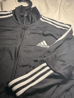Klassisk Adidas zip-up träningsjacka i nyskick – Storlek M - Klassisk Adidas-jacka i nyskick! Säljer en stilren svart träningsjacka från Adidas med de ikoniska tre ränderna längs ärmarna och broderad logotyp på bröstet. En tidlös modell som passar lika bra till gymmet som till vardags. Mycket gott skick, inga slitage, hål eller fläckar. Dragkedjan fungerar felfritt. Finns att hämta i Göteborg eller kan skickas mot fraktkostnad. Tveka inte att höra av dig om du har frågor! 