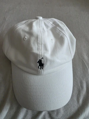 Vit keps från Polo Ralph Lauren - Snygg vit keps från Polo Ralph Lauren med klassisk böjd skärm och broderad svart logga med polospelare framtill. Justerbar rem baktill för perfekt passform. 