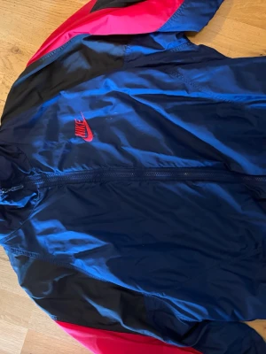 Nike vintage vindjacka - Snygg vindjacka från Nike i marinblått med röda och svarta detaljer. Köpte denna i en vintage butik i New York och knappt används sedan dess. Skicket är som den var när jag köpte den men den är som sagt köpt i en vintage butik. Strl XL men funkar absolut som L.