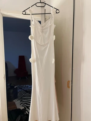 Vit långklänning med rosettdetaljer - Elegant vit långklänning med smala axelband och dekorativa rosetter längs banden. Klänningen har en mjuk drapering vid bysten och en figurnära passform som faller snyggt längs kroppen. Perfekt för sommarens alla tillfällen.