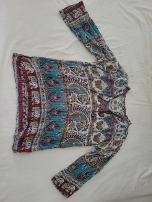 Mönstrad boho-blus med fjärilar - Bohemisk blus med färgstarkt mönster i turkos, vinrött, senapsgult och vitt. Blusen har långa ärmar och ett snöre vid halsringningen. Materialet är tunt och luftigt, perfekt för en avslappnad stil. Dekorerad med fjärilar och blommor för en unik look.