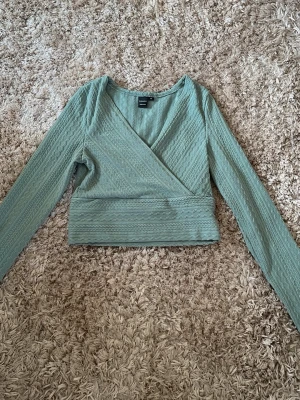 Grön omlottopp från Shein Sky - Snygg ljusgrön omlottopp från Shein Sky med lång ärm och croppad passform. Toppen har ett diskret vågmönster i tyget och bred midjeresår som ger en smickrande siluett. Perfekt att styla med höga jeans eller kjol.