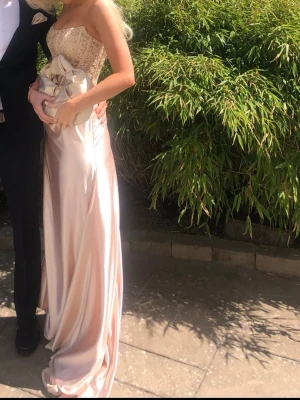 Champagnefärgad balklänning med korsett - Elegant långklänning i champagnefärg med spetsdetaljer på överdelen och axelbandslös design. Klänningen har en figurnära passform och är tillverkad i glansigt satinliknande material som ger ett lyxigt intryck. Perfekt för bal eller student. Köpt i april 2025 och använd 1 gång.